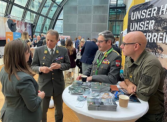 Zahlreiche Menschen am HR Summit. Im Vordergrund drei uniformierte Herren, die mit einer Dame sprechen.