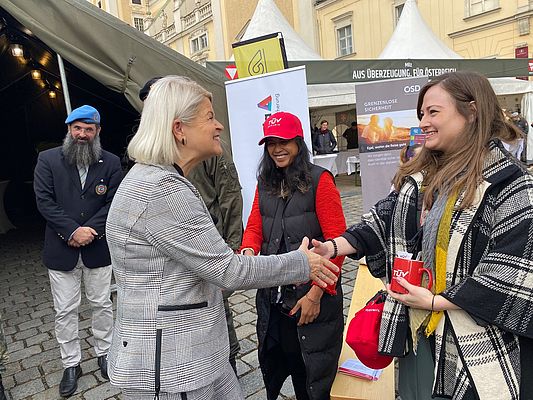 Drei lächelnde Damen. Im Hintergrund zwei Herren.